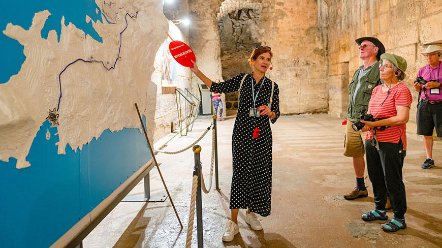 A tour guide in a polka dot dress points to a large wall map while explaining it to three tourists inside a stone room.