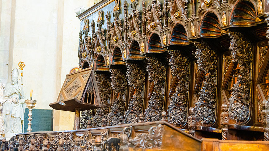 Ornate and intricately carved dark wood choir stalls with a matching lectern inside a stone cathedral next to a bishop statue.