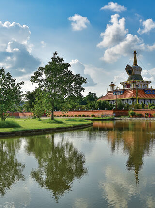 A sacred Buddhist stupa and lush trees reflect in a calm pond at the Buddha's birthplace in Nepal under a bright, cloudy sky.