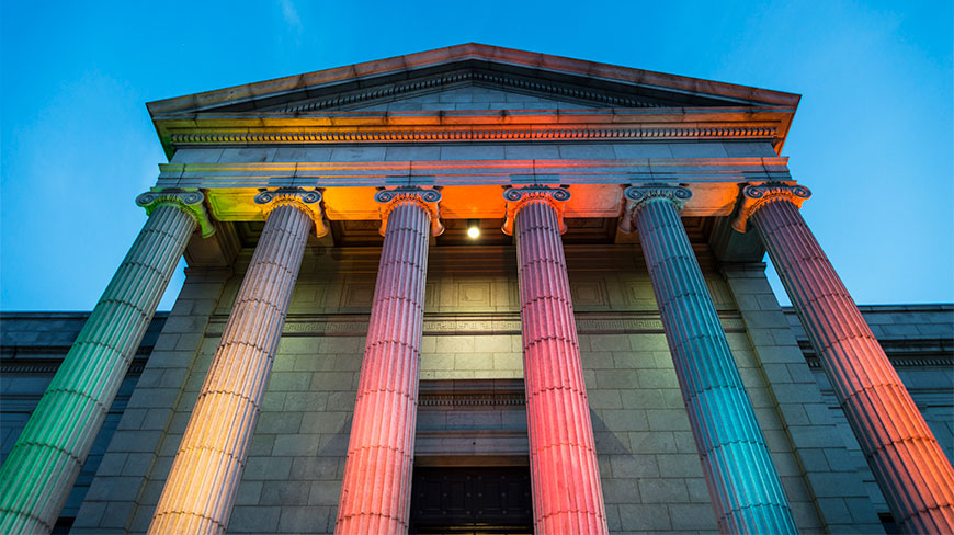 The Minneapolis Institute of Arts facade featuring tall Ionic columns illuminated in rainbow colors at dusk.