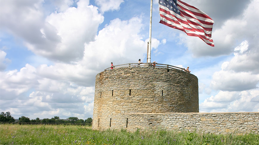 Visitors stand atop the historic round stone tower at Fort Snelling in St. Paul, Minnesota, under a large, waving American flag.