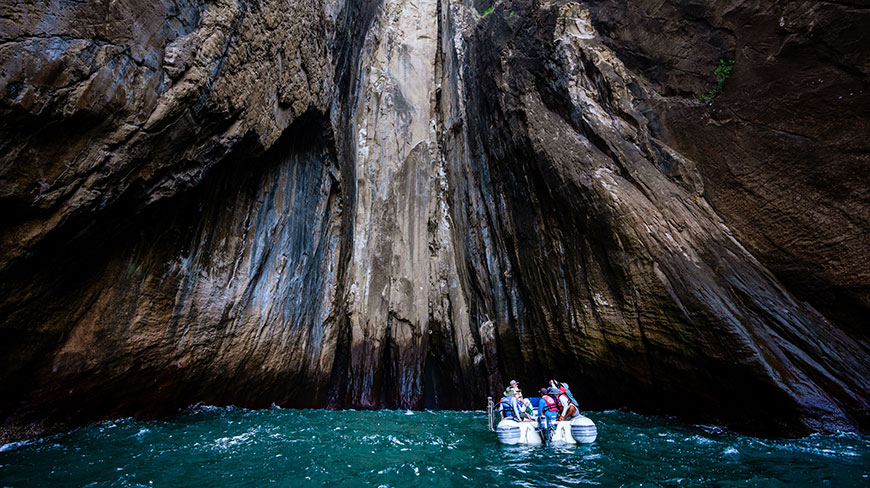 A small inflatable boat with passengers floats on turquoise water at the entrance to a narrow gorge with towering, dark rock walls.