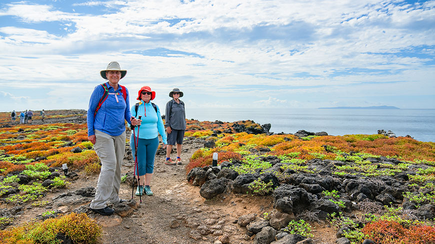 Three women in hiking gear stand on a rocky, coastal trail covered in colorful vegetation, with the ocean in the background under a blue sky.