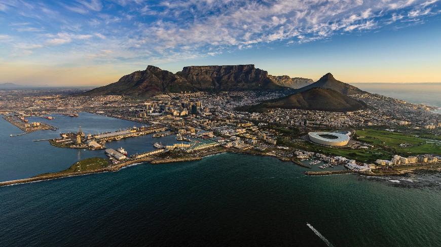 Aerial panoramic view of Cape Town, South Africa, featuring Table Mountain, the city skyline, and the waterfront under a streaked blue sky.