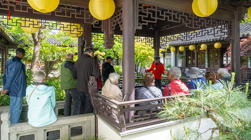 Visitors listen to a guide in a red jacket under a pavilion decorated with yellow lanterns at Lan Su Chinese Garden in Portland.