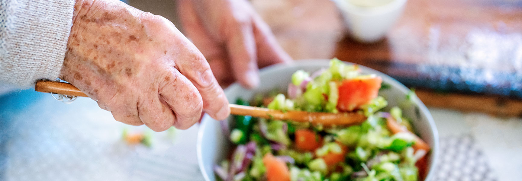 An older person's hands use a wooden spoon to mix a colorful salad in a white bowl.
