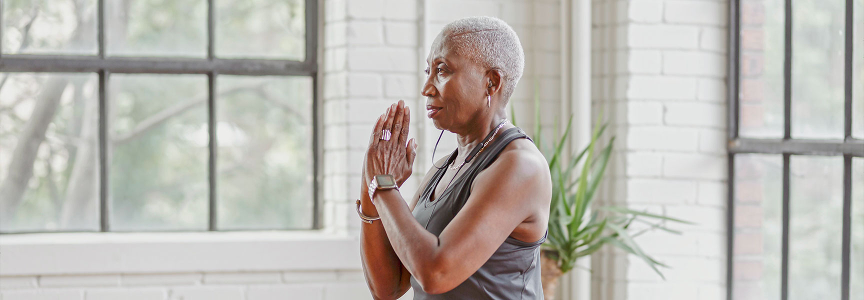 An older Black woman with short gray hair stands in a yoga pose with her hands together in a sunlit room.