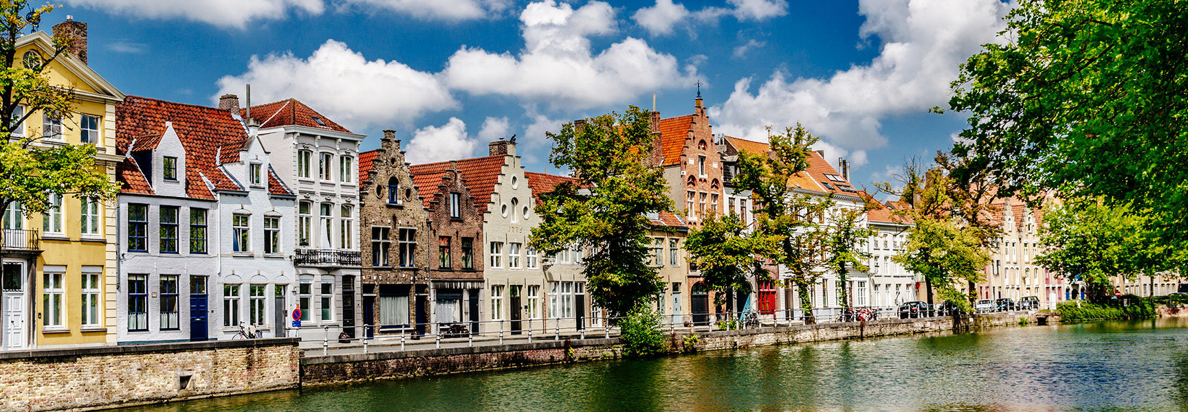 Historic gabled buildings line a calm canal in Bruges, Belgium, reflecting in the water under a bright sky with fluffy white clouds.