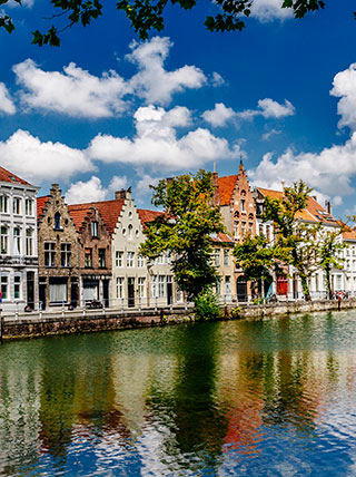 Historic gabled buildings line a calm canal in Bruges, Belgium, reflecting in the water under a bright sky with fluffy white clouds.