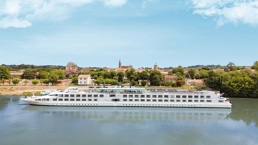 The MS Cyrano de Bergerac cruise ship travels on a calm river with a scenic town and lush greenery in the background.