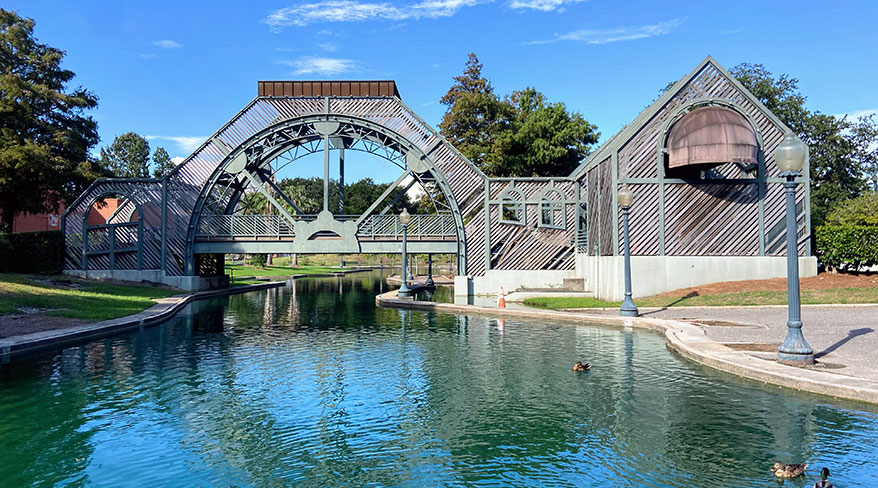 An arched bridge spans a quiet lagoon with ducks swimming in Louis Armstrong Park, New Orleans, under a clear blue sky.