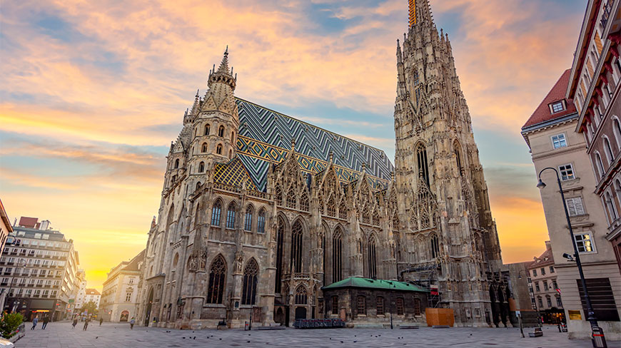 St. Stephen's Cathedral in Vienna with its iconic colorful tiled roof and tall Gothic spire under a glowing sunset sky.