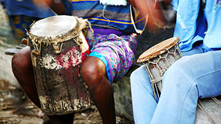 Close-up of two African drummers sitting with traditional talking drums between their knees, with motion blur indicating playing.