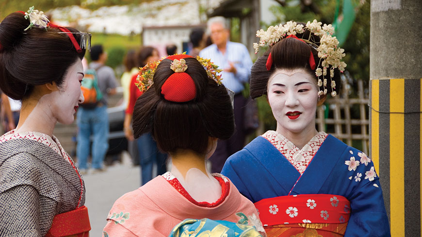 Three Japanese women in traditional geisha attire with white makeup and ornate hairpins stand together on a street.
