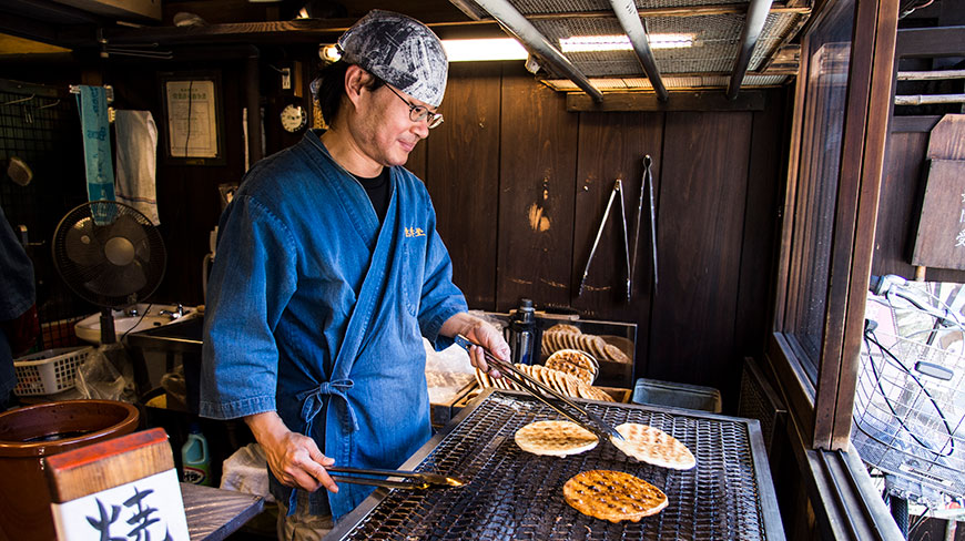A Japanese man in a blue coat grills rice crackers in a small, rustic wooden food stall.