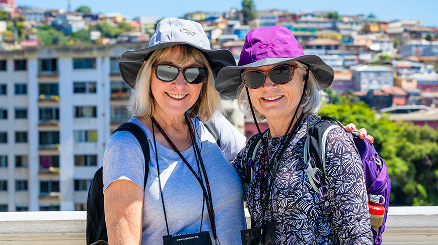 Two women wearing sun hats and sunglasses smile during a Santiago and Valparaiso walking tour with colorful hillside buildings in the background.