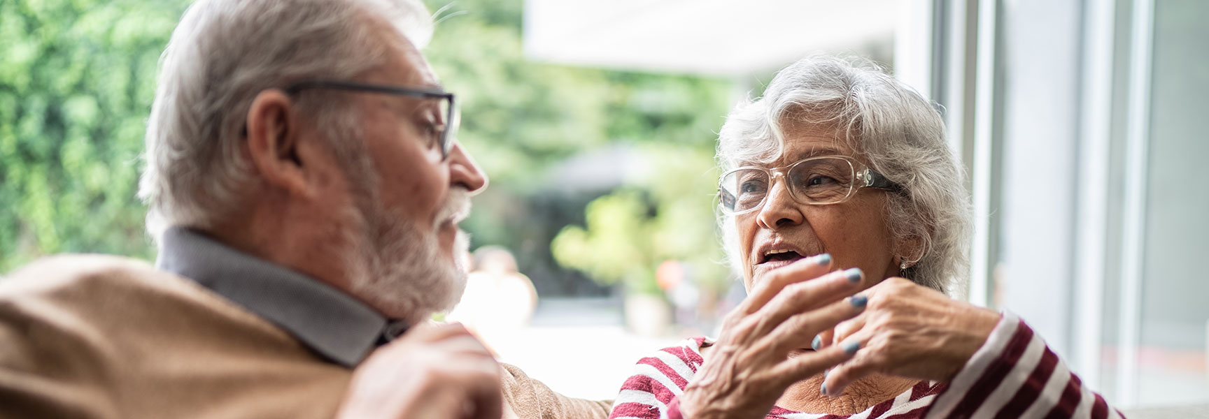 An older woman with glasses gestures as she speaks to a man with a grey beard in front of a bright, leafy window.