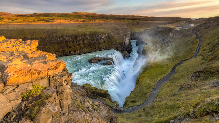 Gullfoss Falls in Iceland cascades into a rugged canyon during sunset, viewed from a high rocky cliff.
