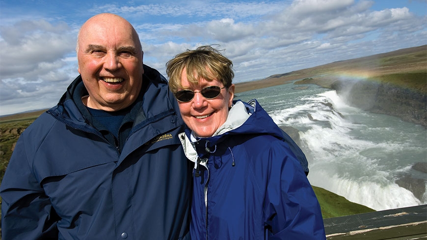 A smiling man and woman in blue jackets pose in front of Gullfoss waterfall with a rainbow visible in the mist.