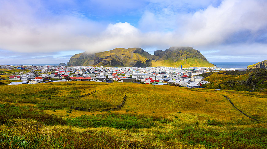 Scenic landscape view of the town of Vestmannaeyjar in the Westman Islands, Iceland, with colorful houses nestled below steep, rugged green mountains.