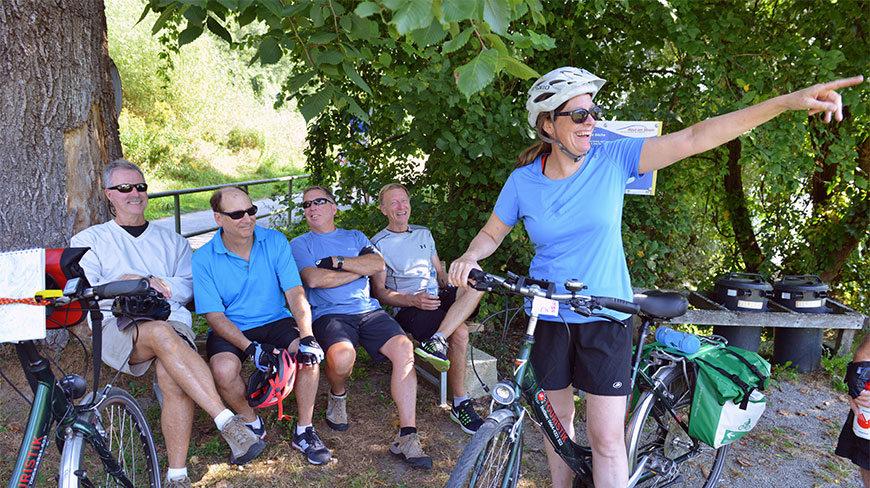 Five cyclists take a break in the shade while biking along the Danube River in Schlögen, with one woman pointing toward the trail.