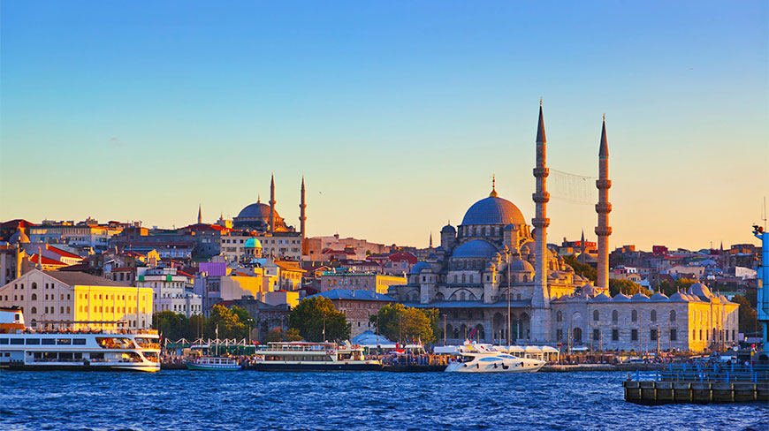 A sunset view of the Istanbul skyline featuring a large mosque and boats along the golden Bosphorus waterfront.