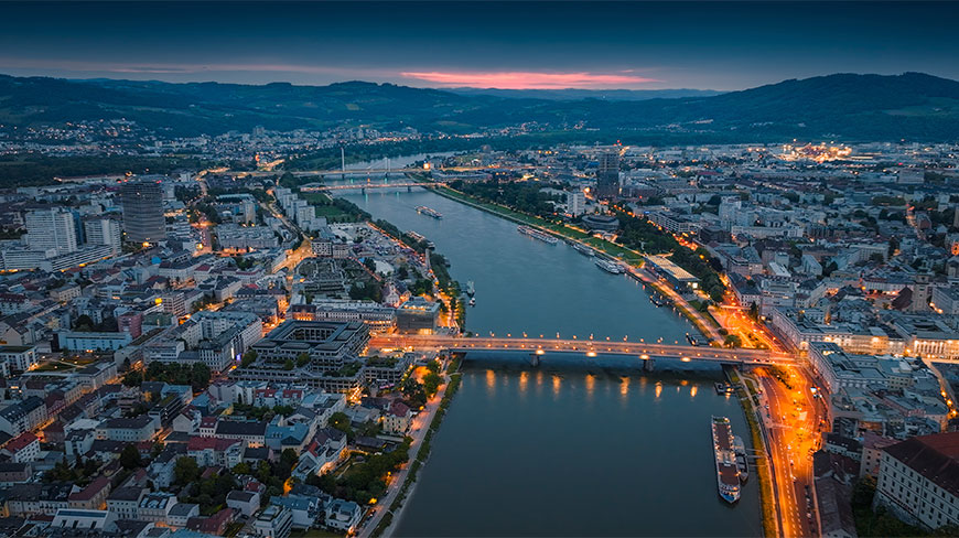 Aerial view of the Danube River flowing through Linz, Austria at dawn, with city lights reflecting on the water and hills in the distance.