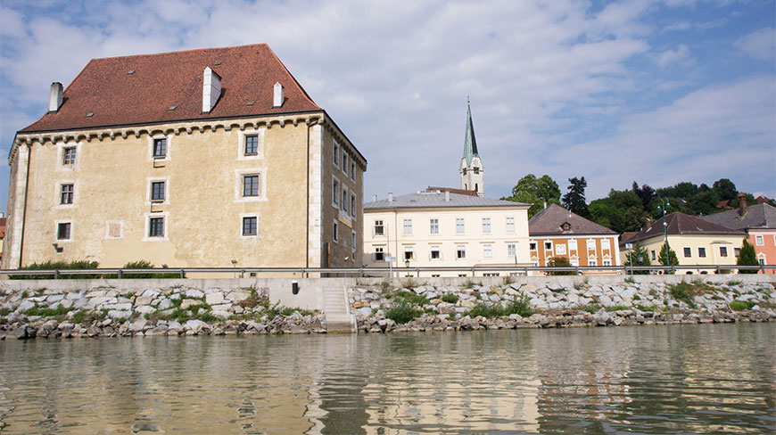 View of Mauthausen Castle and nearby village buildings along the rocky banks of the Danube river under a bright sky.