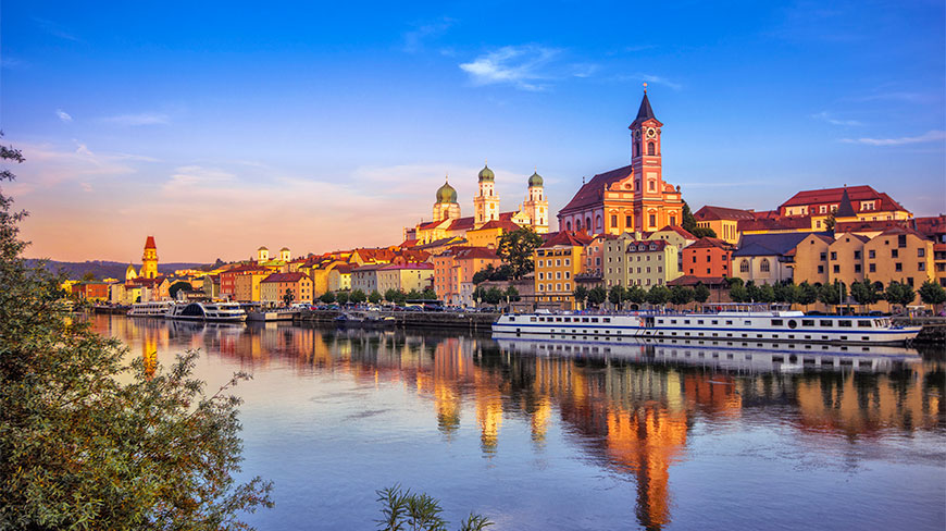 A boat docked on the Danube River in Passau, Germany, with colorful historic buildings and church towers reflected in the water at sunset.