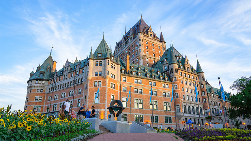 Tourists explore an Old City Quebec walking tour with a view of the grand Fairmont Le Château Frontenac and blooming flowers.