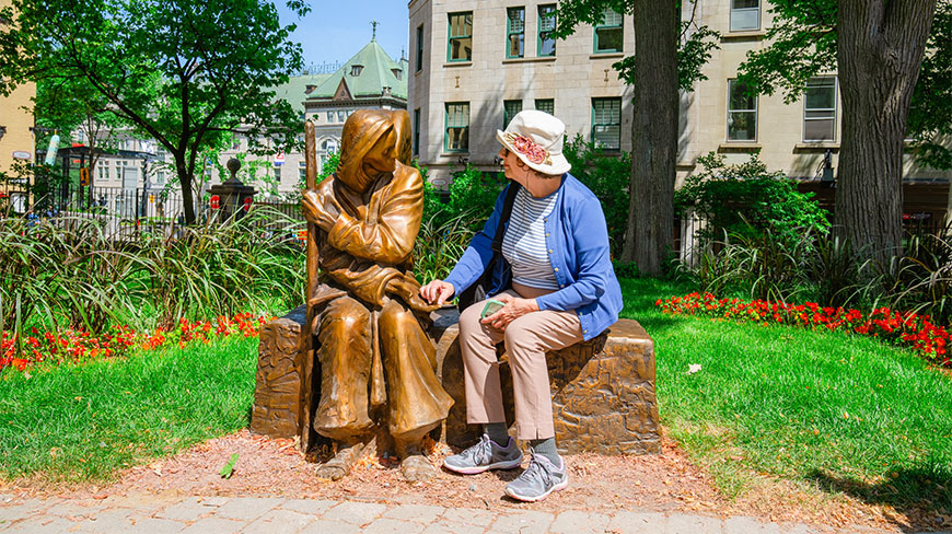 A woman in a blue cardigan and sun hat sits next to a bronze statue during an Old City Quebec walking tour.