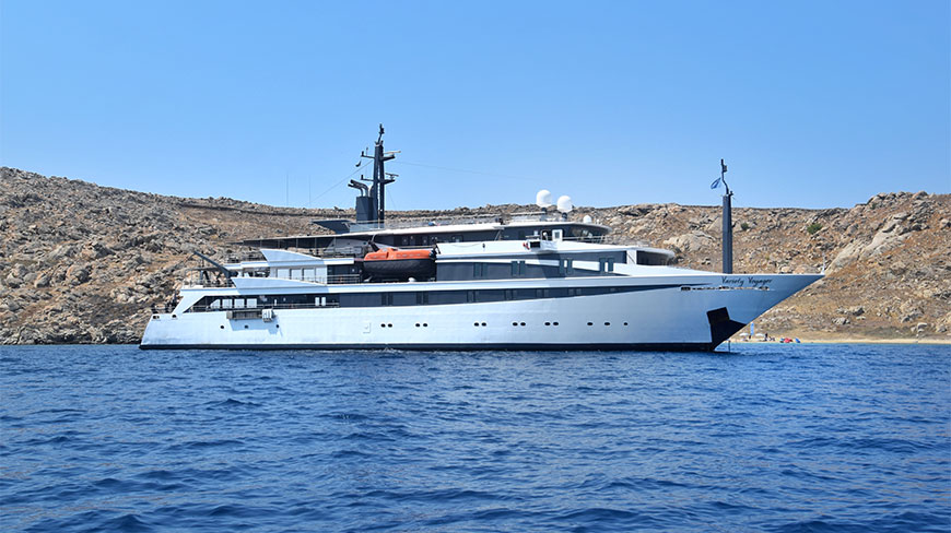 The full exterior of the Variety Voyager ship afloat on calm blue water near a rocky coastline.