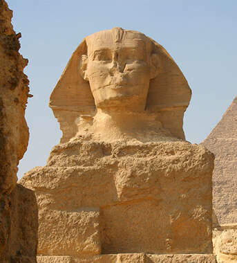 Close-up of the Great Sphinx in Cairo, Egypt, framed by stone ruins with a large pyramid and clear sky in the background.