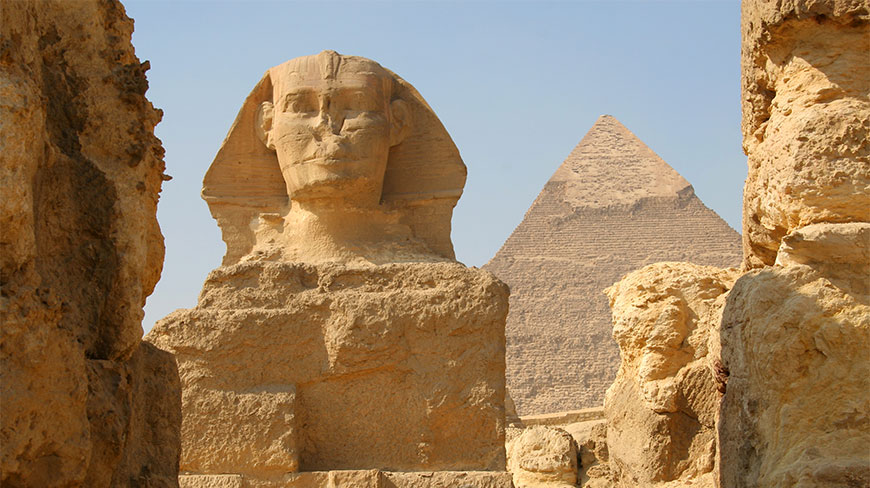 Close-up of the Great Sphinx in Cairo, Egypt, framed by stone ruins with a large pyramid and clear sky in the background.