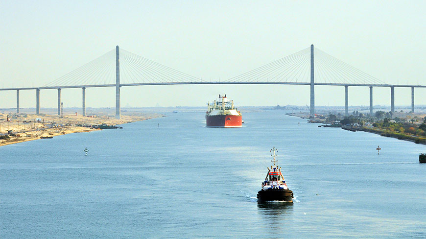 Large cargo ships navigate the Suez Canal under the towering cable-stayed Mubarak Peace Bridge against a clear sky.