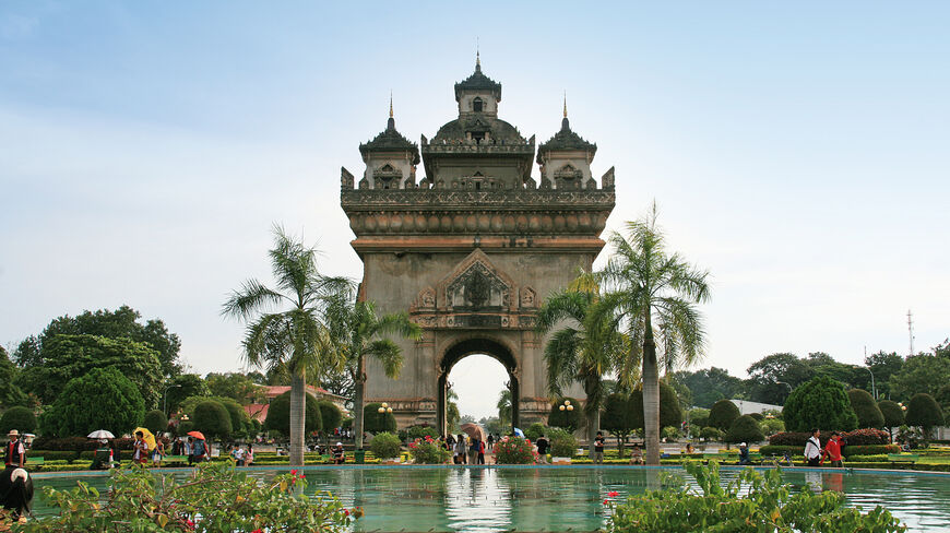 The Vientiane Arc de triomphe stands behind a tranquil reflecting pool and palm trees in a park under a clear blue sky.