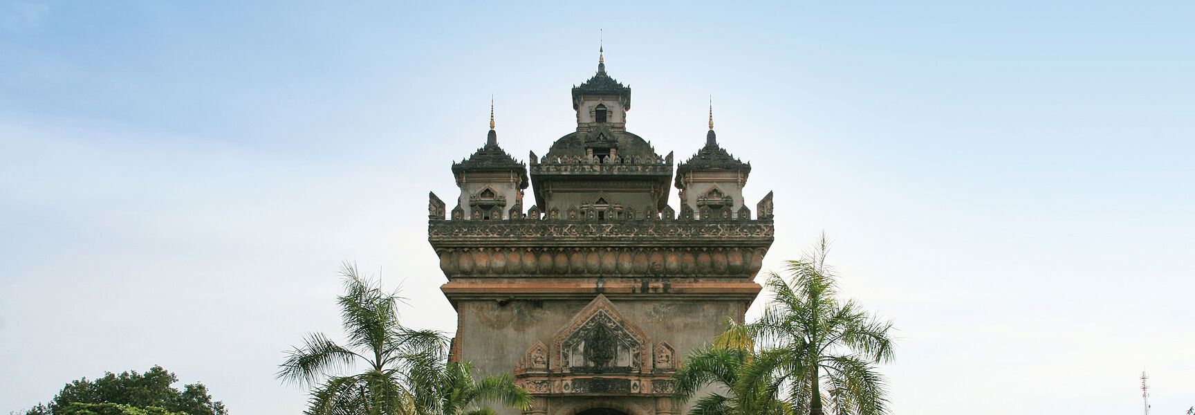 The Vientiane Arc de triomphe stands behind a tranquil reflecting pool and palm trees in a park under a clear blue sky.