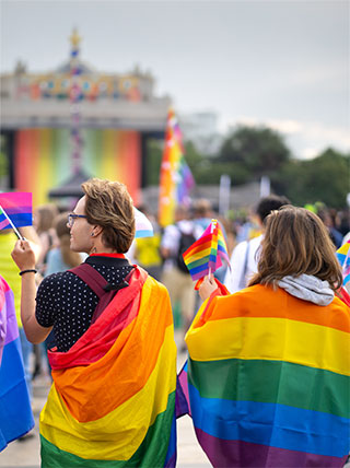 People celebrate at a pride parade, holding rainbow and bisexual flags while draped in colorful pride flag capes.