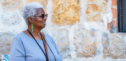 A woman with grey hair and sunglasses stands in front of a stone wall during a walking tour in Oaxaca.