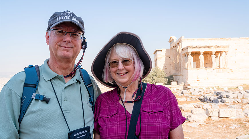 A smiling couple on a morning walking tour of the Athens Acropolis poses in front of the ancient Erechtheion temple ruins.