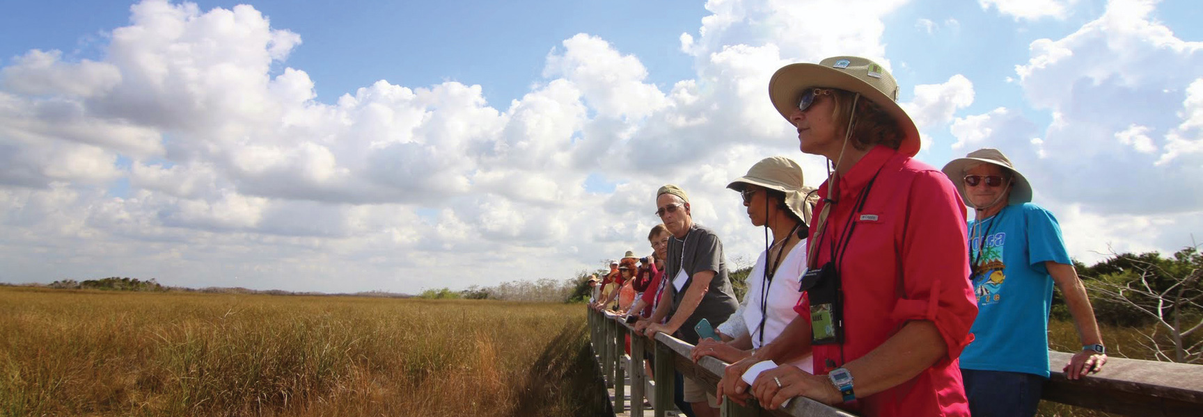A group of people on a boardwalk look out over the vast sawgrass of the Florida Everglades under a cloudy sky.