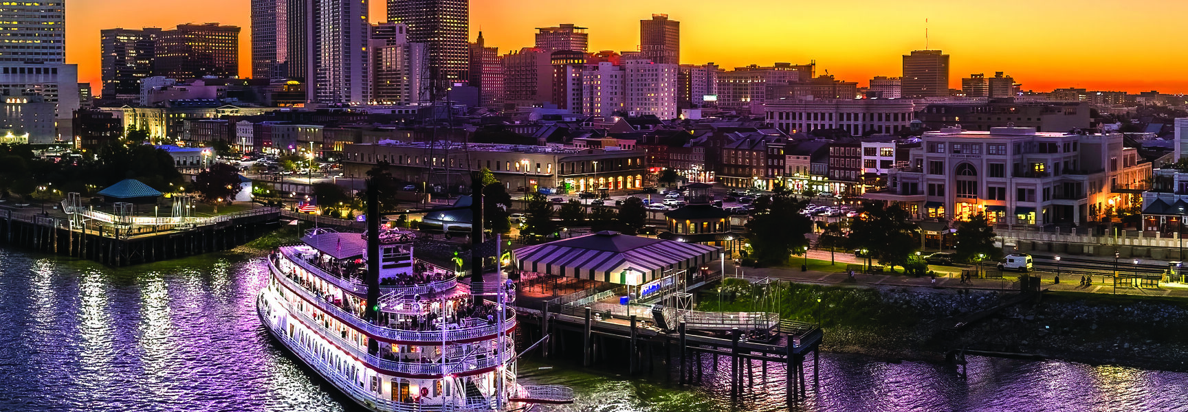 A lit steamboat docked on the Mississippi River with the New Orleans skyline silhouetted against a vibrant orange sunset.