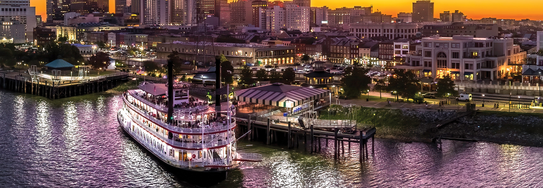 A classic paddle-steamer docks on the riverfront in New Orleans, Louisiana as the sun sets behind the illuminated city skyline.
