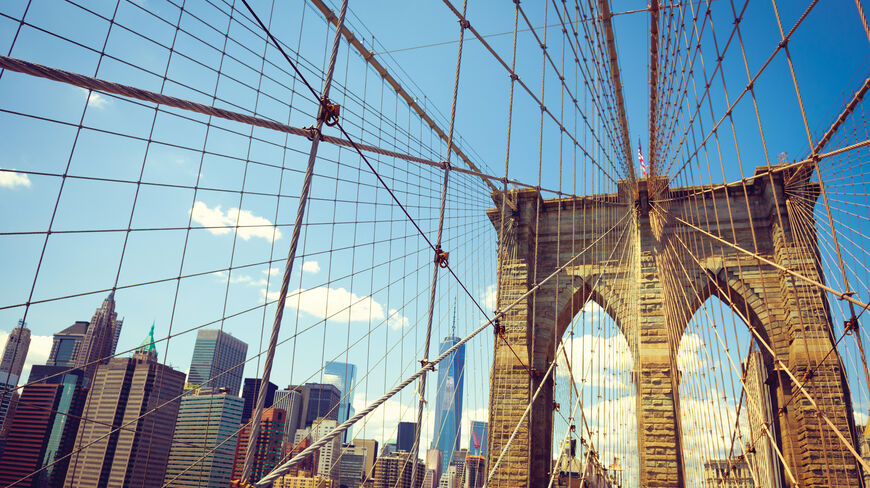 The Brooklyn Bridge's suspension cables and stone tower frame a view of the New York City skyline under a bright blue sky.