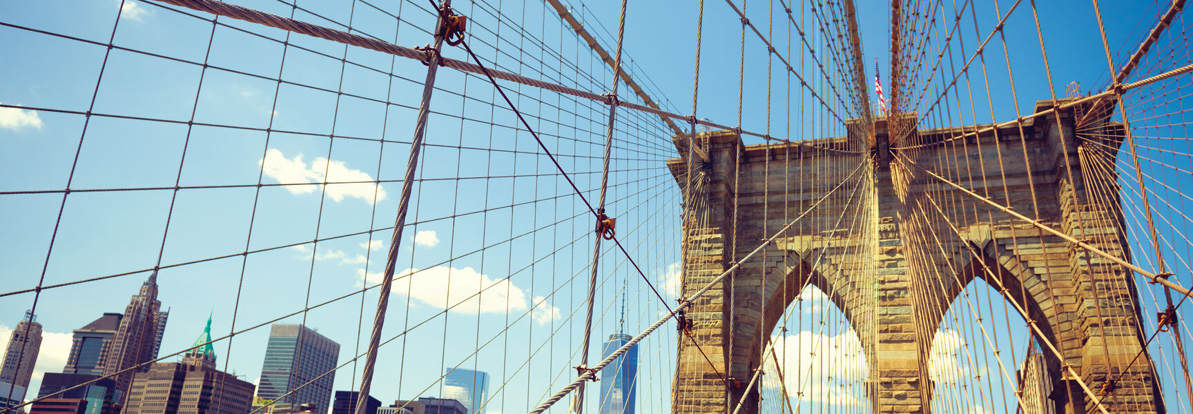 The Brooklyn Bridge's suspension cables and stone tower frame a view of the New York City skyline under a bright blue sky.