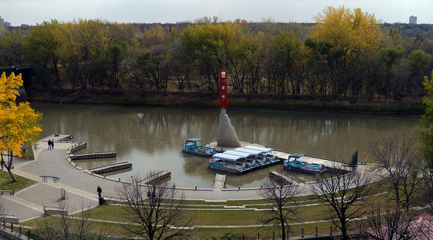 An aerial view of The Forks Historic Port in Winnipeg, Manitoba, showing boats docked along the river surrounded by autumn foliage.