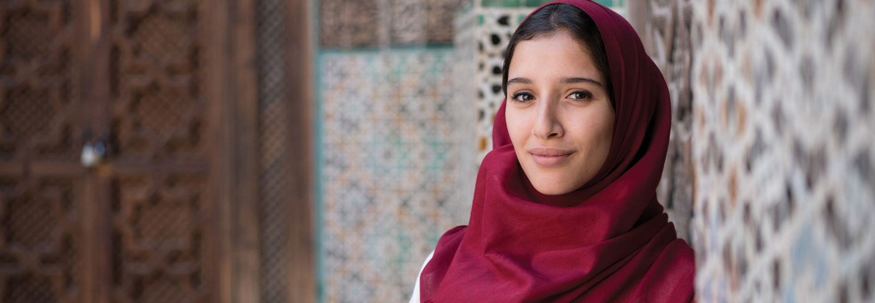 A young woman in a red headscarf smiles gently in front of a traditional mosaic tiled wall in Morocco.