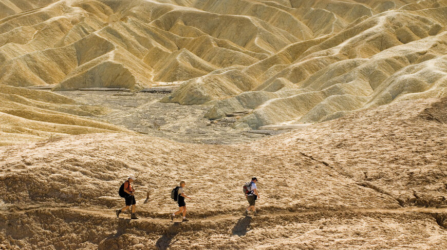 Three people hiking along a desert trail in Death Valley National Park, surrounded by unique, wavy yellow rock formations under bright sunlight.