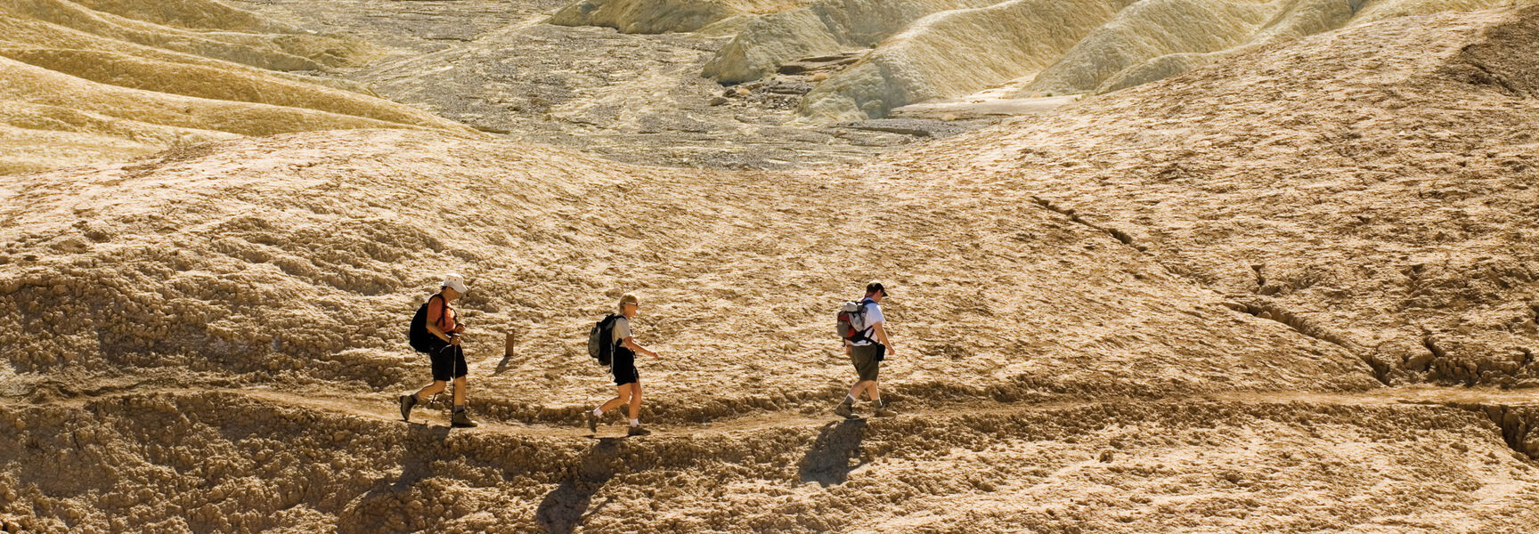 Three people hiking along a desert trail in Death Valley National Park, surrounded by unique, wavy yellow rock formations under bright sunlight.