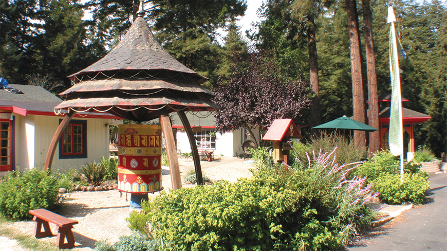 A colorful Buddhist prayer wheel stands in a tranquil garden surrounded by trees and buildings at a retreat in California.